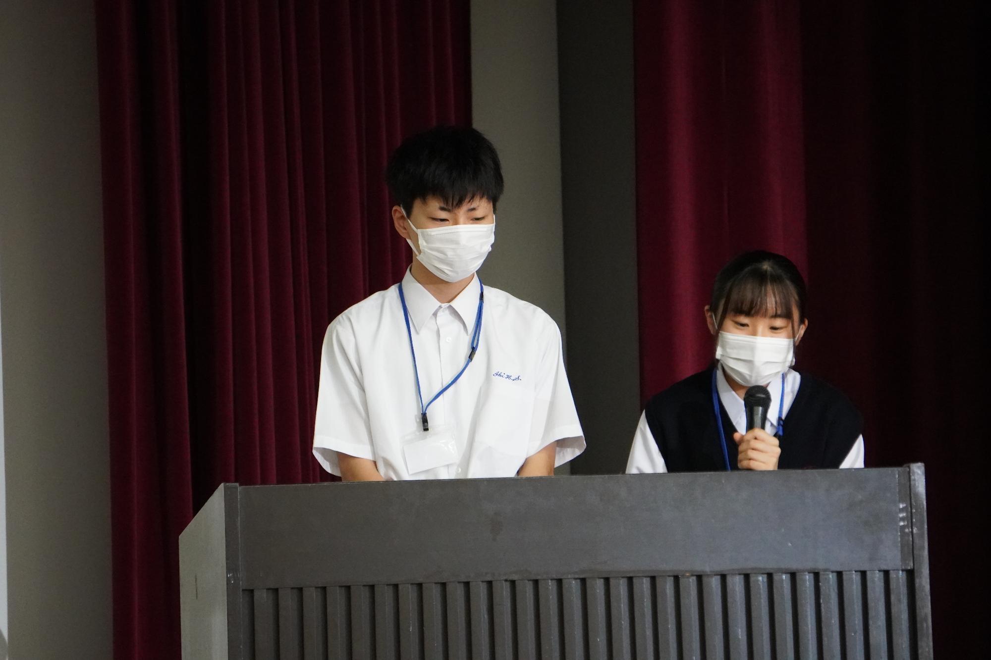 演台の前に男子学生と女子学生が立ち、女子学生がマイクを持ち話をしている写真