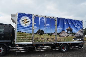 トラック側面にかやぶき屋根のある建物が並び、広い草地が広がっている風景写真のラッピングが施されたラッピングトラックの写真